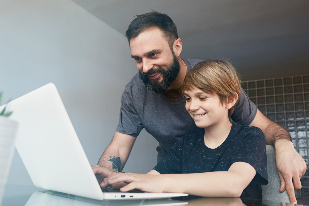 Cheerful young father with cute son using laptop computer while sitting on the table in the living room.